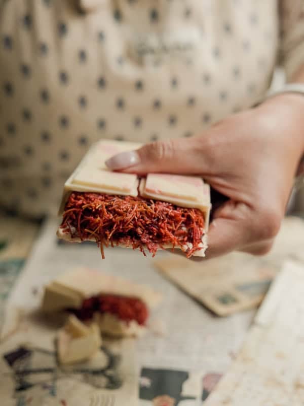 Viral Dubai Chocolate Bar with Strawberry Filling (Vegan). 2 Close-up of a person holding a chocolate bar filled with kataifi. The chocolate is broken, revealing the bright red saffron inside. Newspaper pages are used as the surface beneath the chocolatiers hand.
