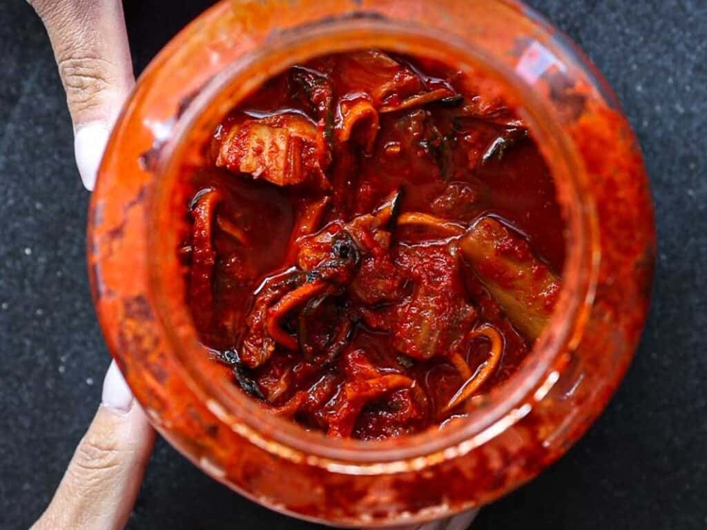 Healthy & Easy Korean Vegan Kimchi Fried Rice Recipe. 4 Open jar filled with red, spicy kimchi, showcasing pieces of fermented vegetables. Two hands hold the jar on a dark surface, highlighting the rich, vibrant colors and textures.