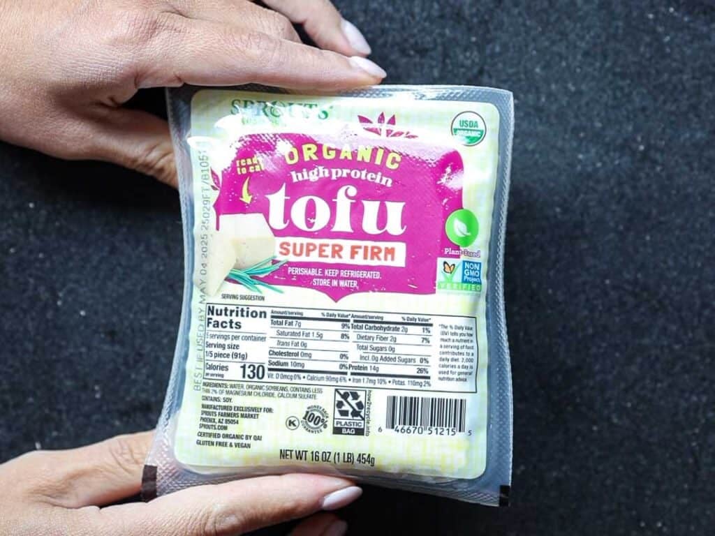 Healthy & Easy Korean Vegan Kimchi Fried Rice Recipe. 6 Two hands hold a package of organic high-protein super firm tofu. The packaging includes nutrition facts, a USDA organic label, and recycling symbols. The background is a textured black surface.