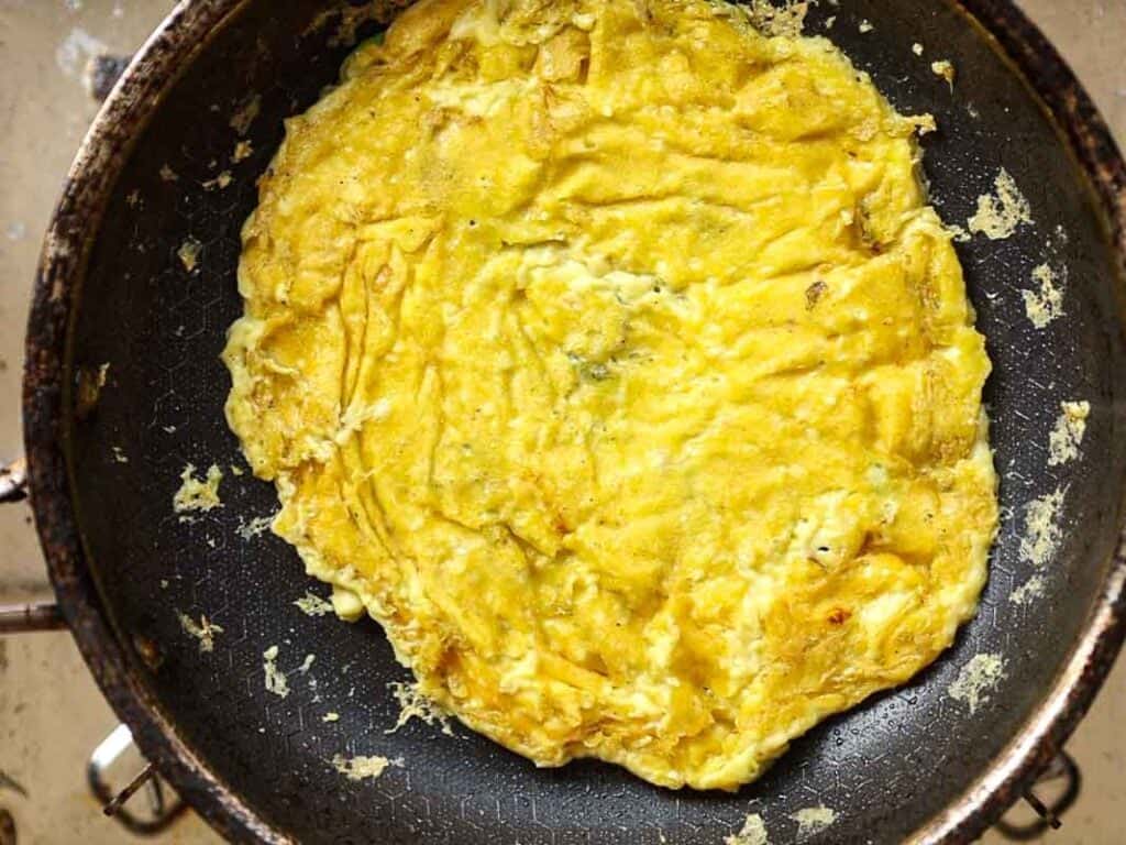 Healthy & Easy Korean Vegan Kimchi Fried Rice Recipe. 9 A golden-brown omelette cooking in a black frying pan. The surface of the omelette is slightly crispy with visible folds and edges. The pan is on a stovetop with bits of egg scattered around.