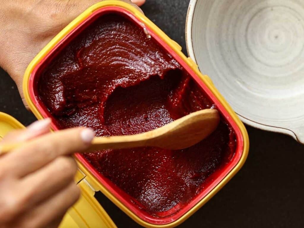 Healthy & Easy Korean Vegan Kimchi Fried Rice Recipe. 20 A hand holds a wooden spoon scooping dark red paste from a rectangular yellow container. A light-colored bowl is partially visible beside the container. The paste has a smooth texture.