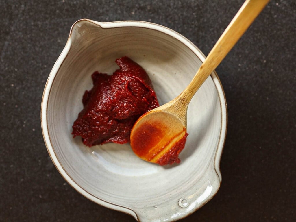 Healthy & Easy Korean Vegan Kimchi Fried Rice Recipe. 21 A ceramic bowl with a scoop of dark red gochujang paste is placed against a dark background. A wooden spoon rests in the bowl, partially dipped in the paste.