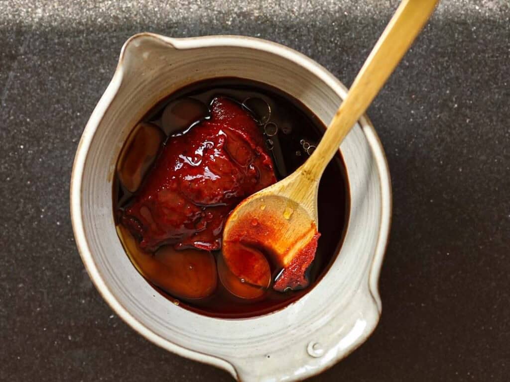 Healthy & Easy Korean Vegan Kimchi Fried Rice Recipe. 22 A ceramic bowl containing gochujang sauce. A wooden spoon rests inside, covered in the sauce. The bowl is placed on a dark, textured surface.