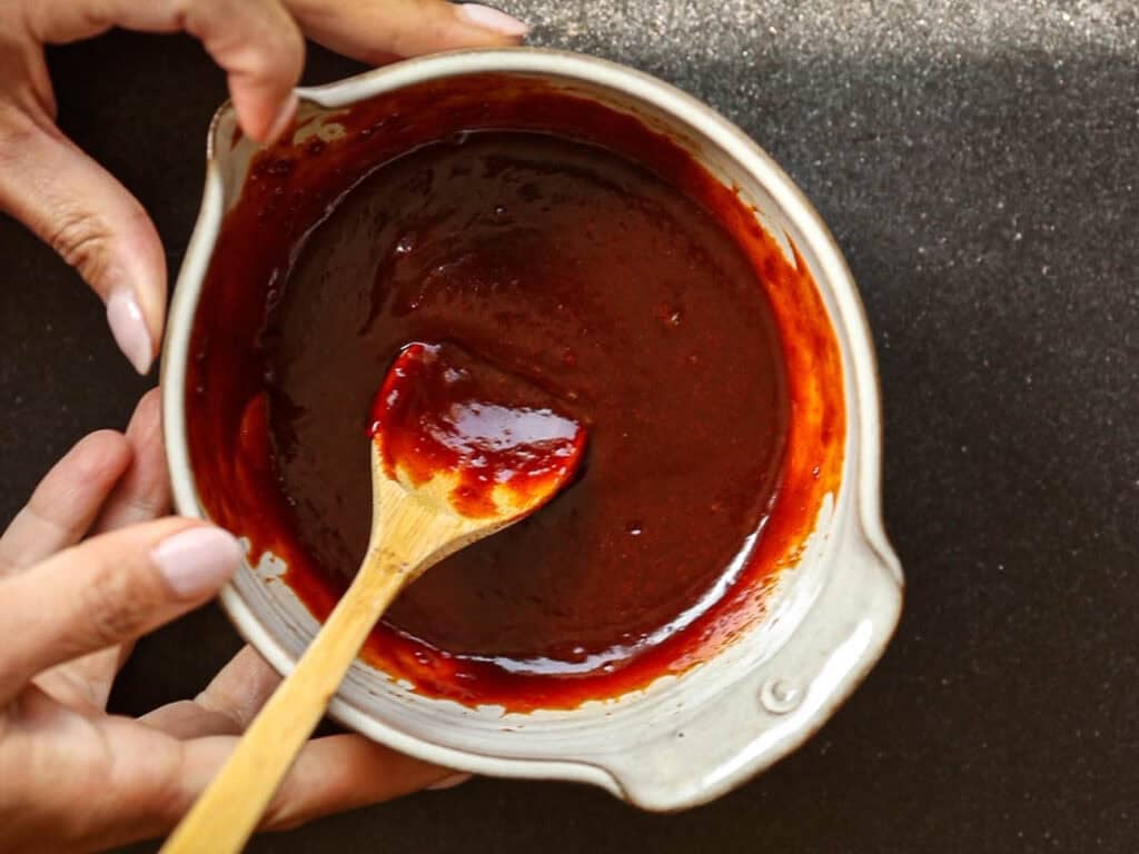 Healthy & Easy Korean Vegan Kimchi Fried Rice Recipe. 8 Hands hold a bowl of dark red gochujang sauce, with a small wooden spoon resting inside. The background is a dark textured surface.