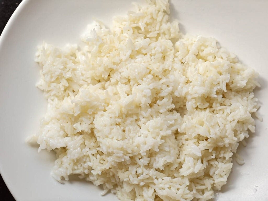 Healthy & Easy Korean Vegan Kimchi Fried Rice Recipe. 5 A white plate filled with cooked white rice, with individual grains visible and slightly fluffy in texture. The plate is positioned on a dark surface.