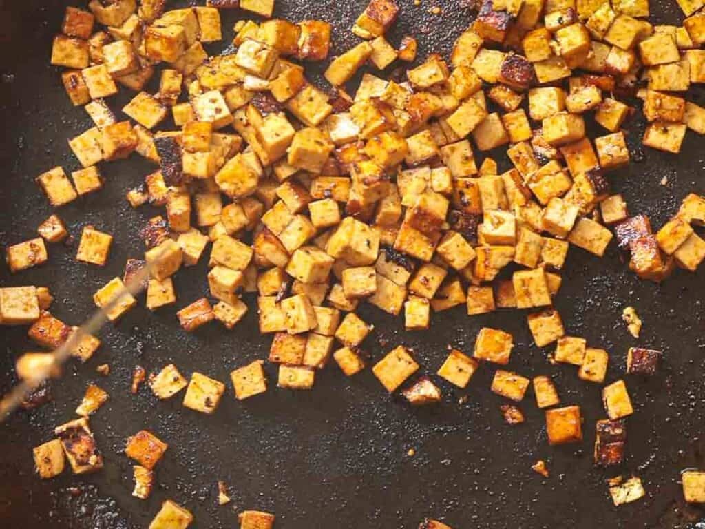 Cubes of tofu are being pan-fried to a golden brown on a black skillet. The tofu pieces are evenly scattered, showcasing a crispy texture with lightly charred edges.