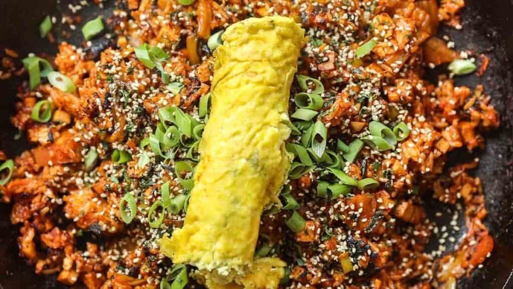 Healthy & Easy Korean Vegan Kimchi Fried Rice Recipe. 1 A close-up of vegan kimchi fried rice topped with a rolled omelette. The dish is garnished with sliced green onions and sesame seeds. The rice appears spicy, with a rich red hue, and is served on a dark plate.