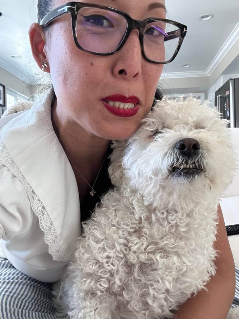 These pics of Lulu are GUARANTEED to make you smile!!! 11 A woman with glasses and red lipstick is taking a selfie with a curly-haired white dog. They are indoors, and the dogs eyes are closed. The woman is wearing a white top and striped pants.