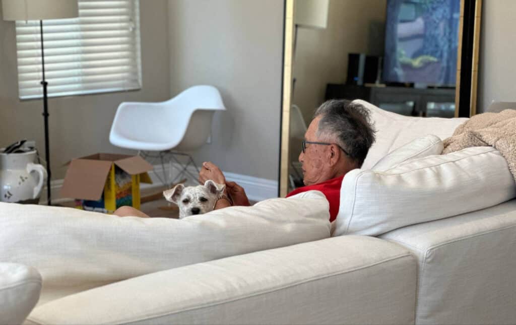 An elderly man sits on a white sofa with a small dog resting beside him. A cardboard box is open on the floor. A white modern chair and a TV can be seen in the background. The man looks relaxed and is barefoot.