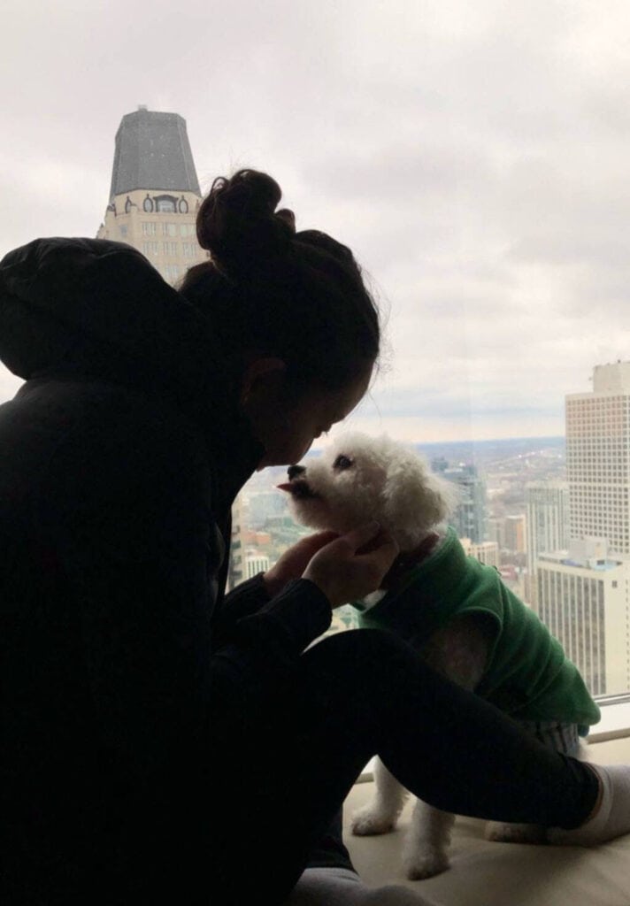 These pics of Lulu are GUARANTEED to make you smile!!! 2 Joanne, in a dark jacket, sits by a window, holding Rudy, a small white dog wearing a green sweater. They face each other affectionately against a cityscape backdrop with tall buildings and a cloudy sky.
