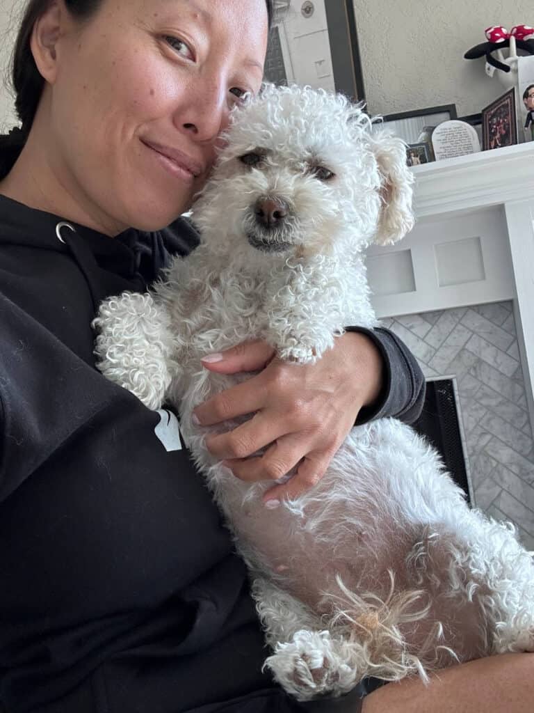 These pics of Lulu are GUARANTEED to make you smile!!! 12 A person wearing a black hoodie gently holds a small, curly-haired white dog in their arms. The dog looks content with its eyes half-closed. They are sitting in a cozy room with a fireplace and picture frames in the background.