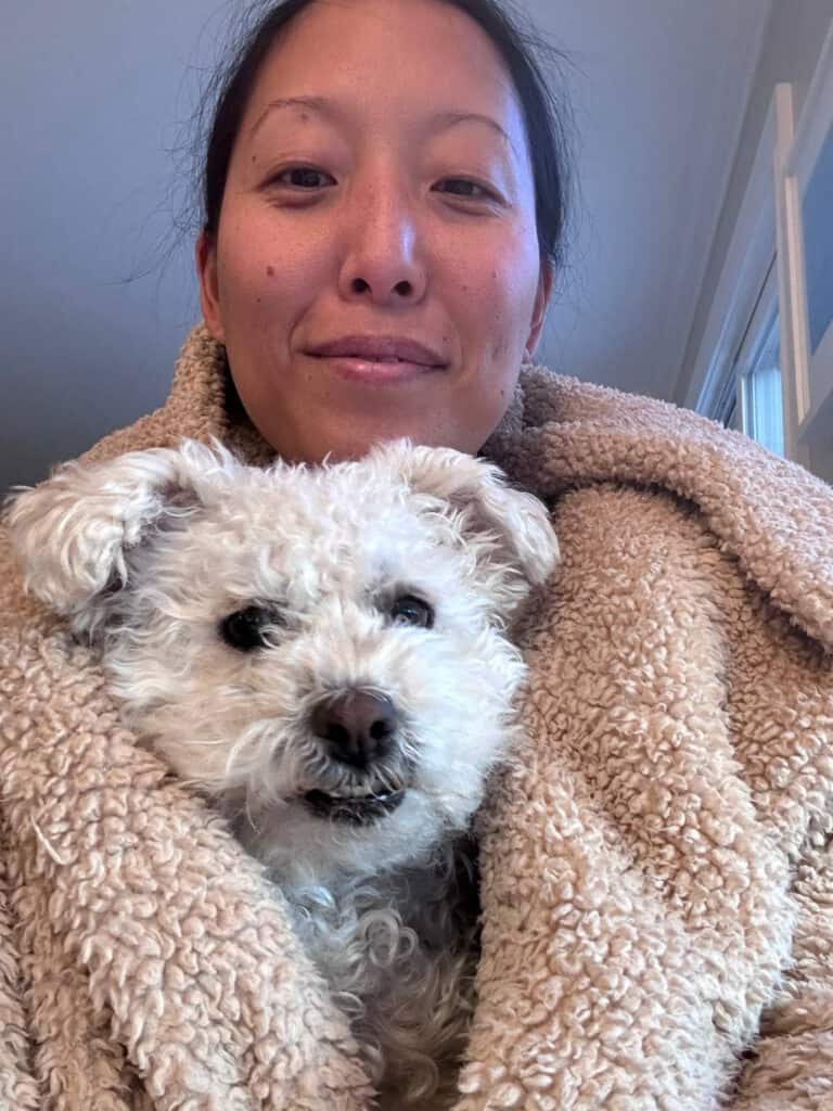 Joanne wrapped in a fluffy beige blanket smiles at the camera with Lulu, a small, curly-haired white dog cozily nestled within the blanket. They are indoors with a light background.