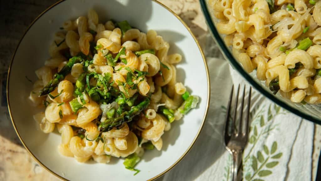 30-Minute Delicious Asparagus Pasta Recipe (Vegan). 14 A bowl of creamy asparagus pasta recipe with asparagus and herbs is served on a table. Another bowl of the same dish is nearby, accompanied by a fork resting on a floral-patterned cloth. Bright natural light highlights the foods textures and colors.