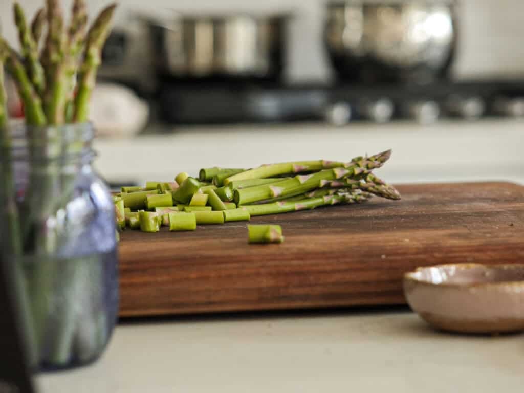 30-Minute Delicious Asparagus Pasta Recipe (Vegan). 3 A cutting board with chopped asparagus spears on a kitchen counter. A jar with whole asparagus is visible on the left, and there are kitchen appliances blurred in the background.