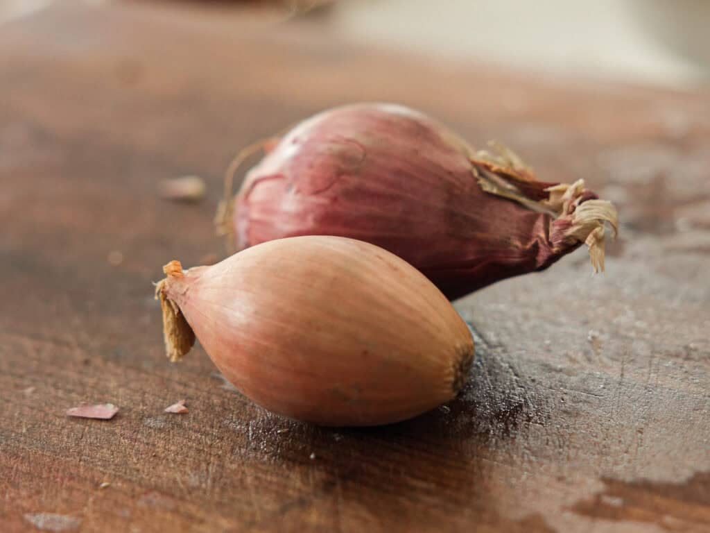 The BEST Sweet Green Pea Risotto Recipe (Vegan). 6 Two shallots, one with a reddish-brown skin and the other with a pinkish hue, are placed on a wooden cutting board. The focus is on their textures and natural sheen.