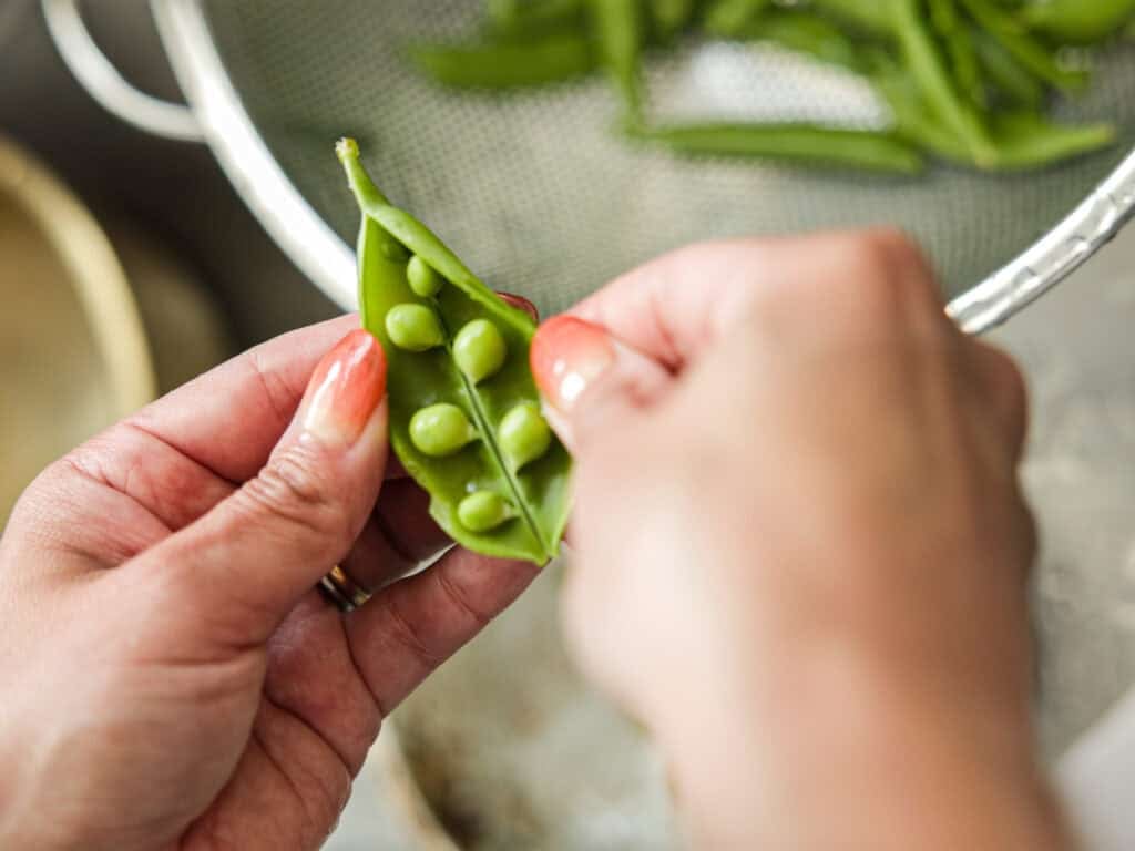 30-Minute Delicious Asparagus Pasta Recipe (Vegan). 4 Hands opening a green pea pod to reveal several peas inside. A colander filled with more pea pods is visible in the background.