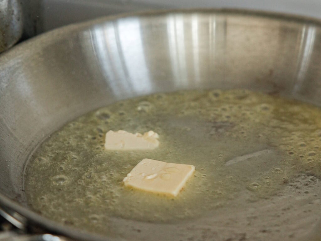 Two pieces of vegan butter melting in a hot stainless steel frying pan, creating a bubbling and sizzling effect on the surface. The butter is beginning to turn golden brown as it melts.
