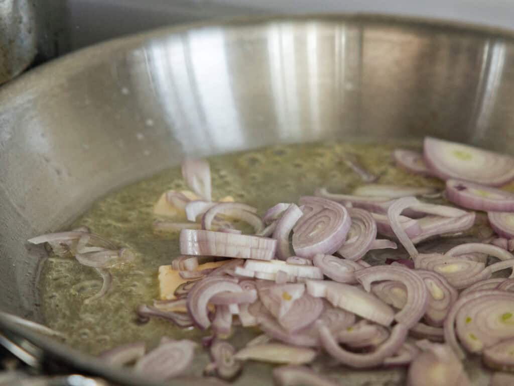 Chopped shallots are sautéing in a stainless steel pan with vegan butter, releasing steam. The pan has a shiny interior, reflecting light. The shallots are sliced thinly, in various sizes, and appear slightly translucent and caramelizing.