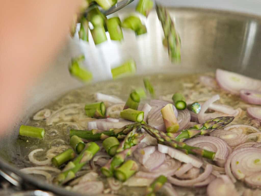 Close-up of a pan with sliced shallots and asparagus being added, sizzling in oil. The thin, circular slices of shallots and chopped asparagus spears suggest the start of a sautéed dish. A hand is slightly blurred, adding ingredients.