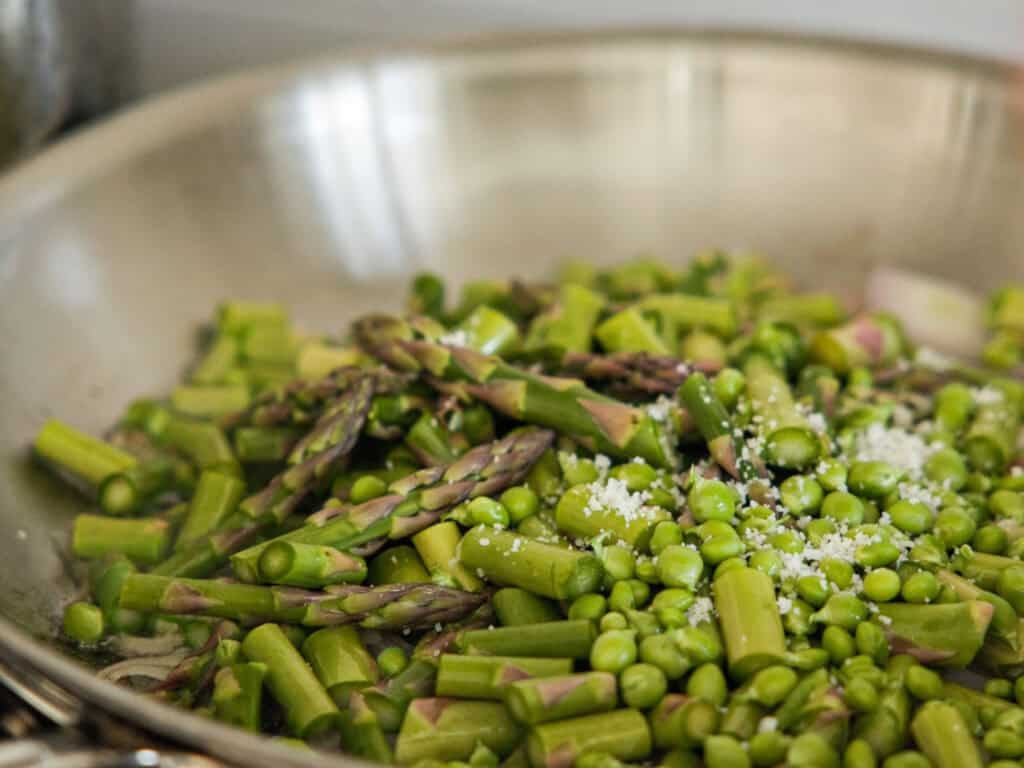 Close-up of a pan filled with chopped green asparagus and peas being cooked. Sea salt is sprinkled on top, adding texture and contrast to the vibrant green vegetables. The stainless steel pan reflects light, enhancing the fresh appearance.
