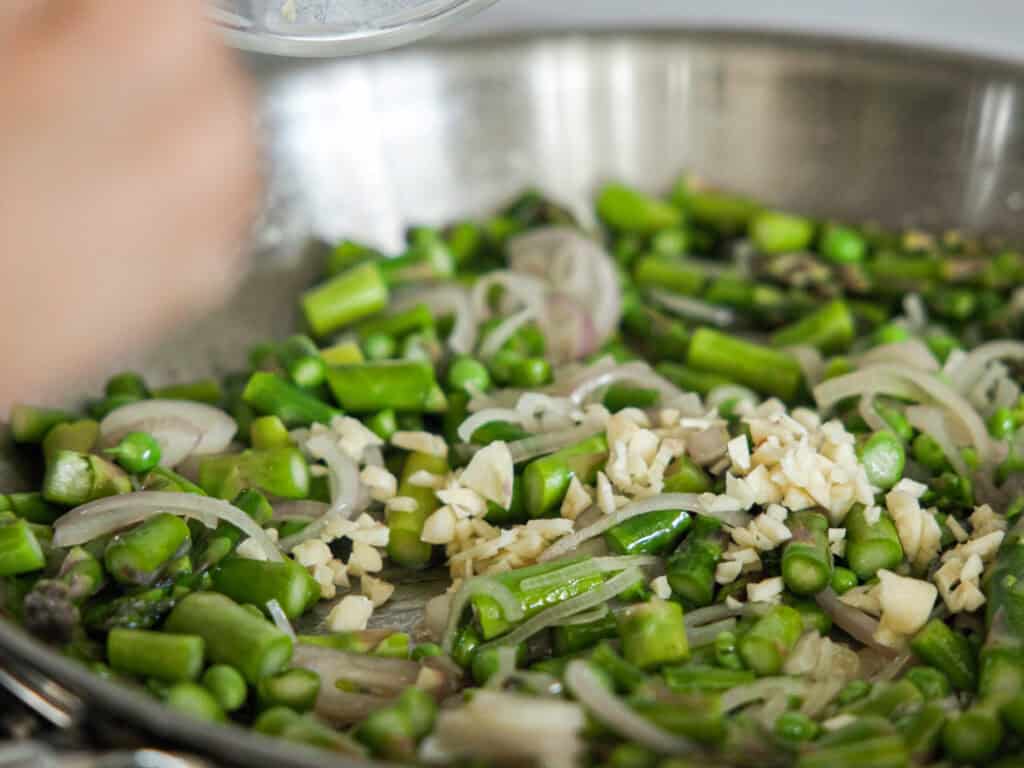 A close-up of a skillet containing chopped green asparagus, sliced onions, and minced garlic being sautéed. A hand is visible in the foreground, adding ingredients. The vegetables appear fresh and vibrant.