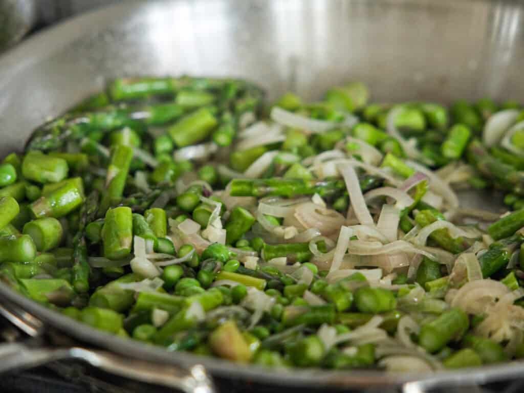 A close-up of a frying pan containing chopped asparagus and sliced onions being sautéed. The vibrant green of the asparagus contrasts with the pale, translucent onions, and the glistening oil suggests they are cooking over heat.