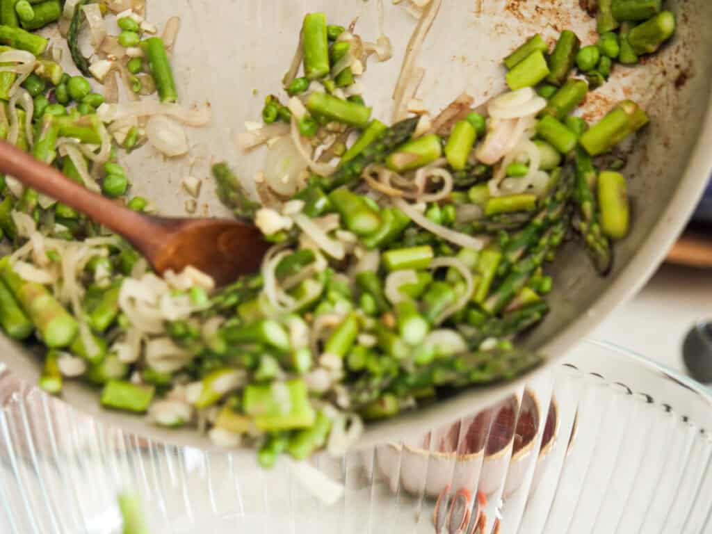 A close-up of sautéed green vegetables being poured from a pan into a dish. The mixture includes asparagus pieces, sliced onions, and peas, stirred with a wooden spoon.