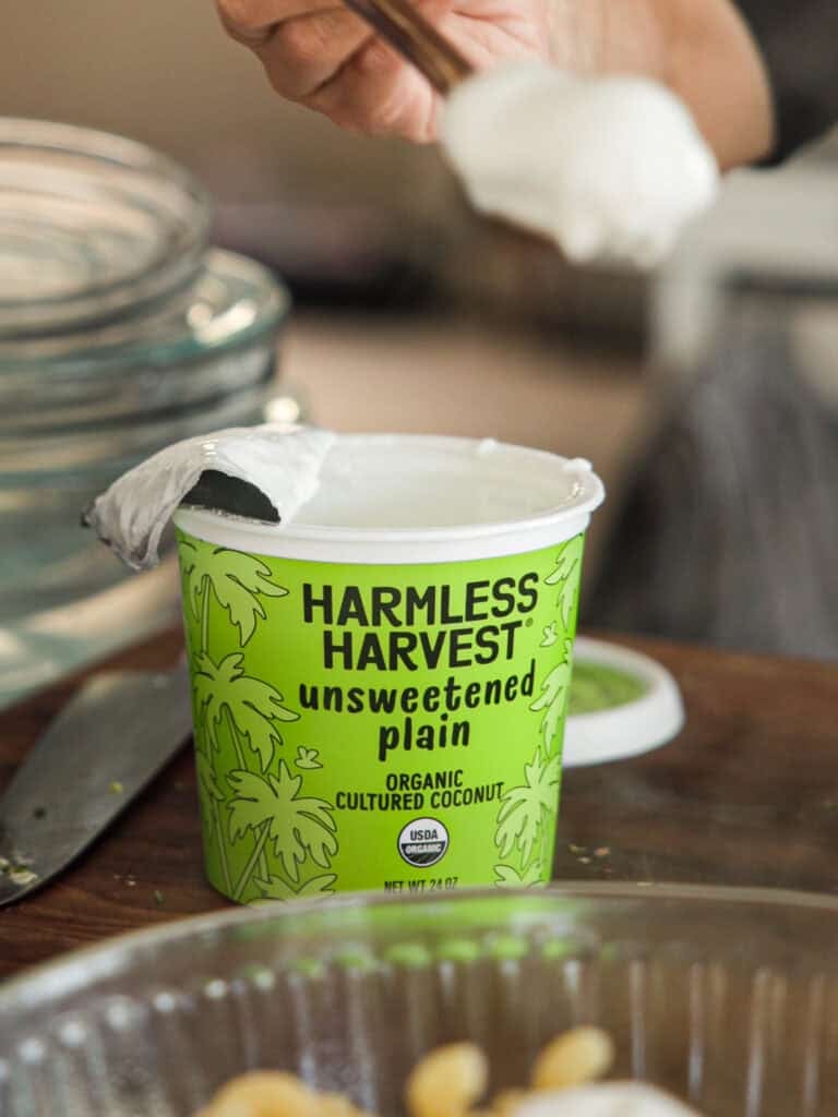 The BEST Sweet Green Pea Risotto Recipe (Vegan). 7 Close-up of a person scooping white yogurt from a green Harmless Harvest container labeled unsweetened plain. The container features palm tree illustrations and text indicating its organic cultured coconut yogurt.