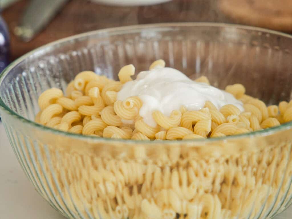 A glass bowl filled with cooked spiral pasta, topped with a dollop of white sauce, likely sour cream or a similar creamy topping, placed on a kitchen counter.