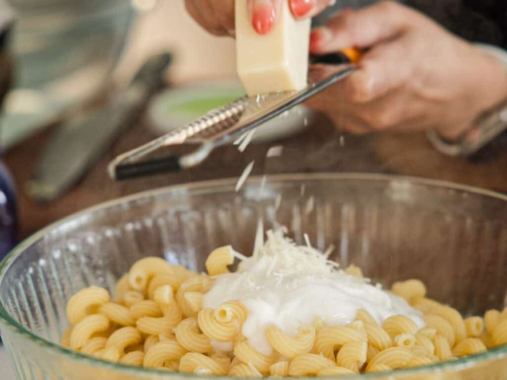 A person grates vegan cheese over a bowl of pasta, topped with a creamy white sauce. The pasta is spiral-shaped and the person has red nail polish. Other kitchen items are blurred in the background.
