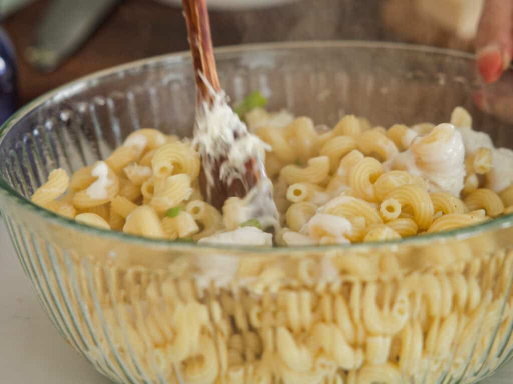 A glass bowl filled with cooked pasta, being mixed with a creamy white sauce using a wooden spoon. Steam is visible rising from the dish, indicating that it is hot.