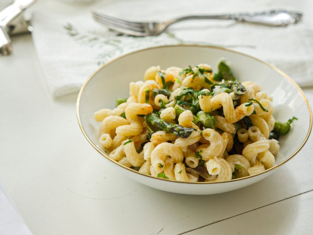 30-Minute Delicious Asparagus Pasta Recipe (Vegan). 18 A bowl of pasta with creamy sauce, asparagus, and herbs, placed on a white table. Silverware and a patterned cloth napkin are placed nearby.