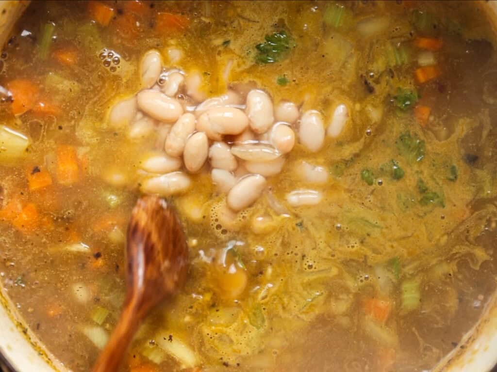 Creamy Cozy Kale White Bean Soup (EASY & Vegan). 5 A wooden spoon stirs a pot of soup with white beans, diced carrots, celery, and herbs in a golden broth.