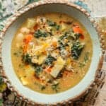 30+ AMAZING VEGAN Holiday Recipes Everyone's gonna Love. 7 A rustic ceramic bowl filled with kale white bean soup featuring carrots, kale, potatoes, beans, and shredded cheese on top, set on a surface decorated with sheet music and colorful illustrations.