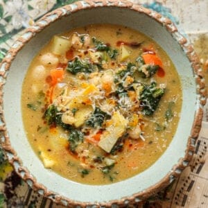 Creamy Cozy Kale White Bean Soup (EASY & Vegan). 27 A rustic ceramic bowl filled with kale white bean soup featuring carrots, kale, potatoes, beans, and shredded cheese on top, set on a surface decorated with sheet music and colorful illustrations.