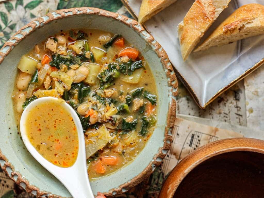 Creamy Cozy Kale White Bean Soup (EASY & Vegan). 3 A bowl of hearty vegetable soup with beans, carrots, greens, and potatoes, topped with a ceramic spoon. Slices of crusty bread sit on a plate nearby. Warm, rustic table setting.