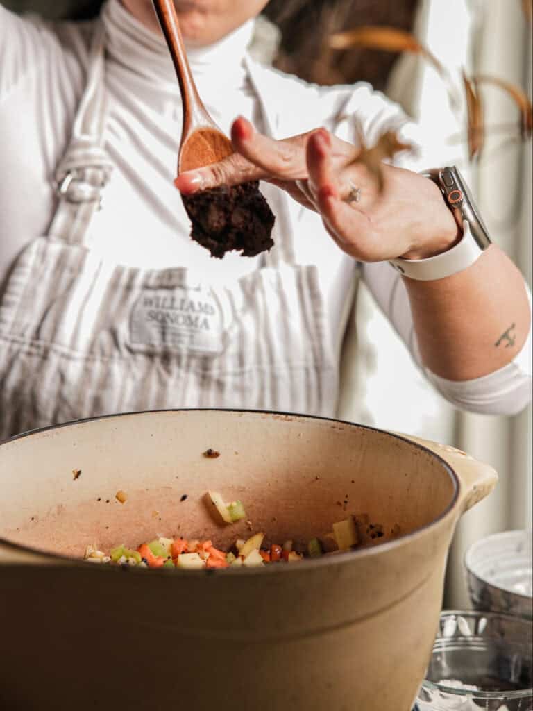 Creamy Cozy Kale White Bean Soup (EASY & Vegan). 12 A person in a white apron adds a spoonful of doenjang to a pot of chopped vegetables, with their other hand using a finger to scrape the spoon. The pot is beige and sits on a kitchen counter.