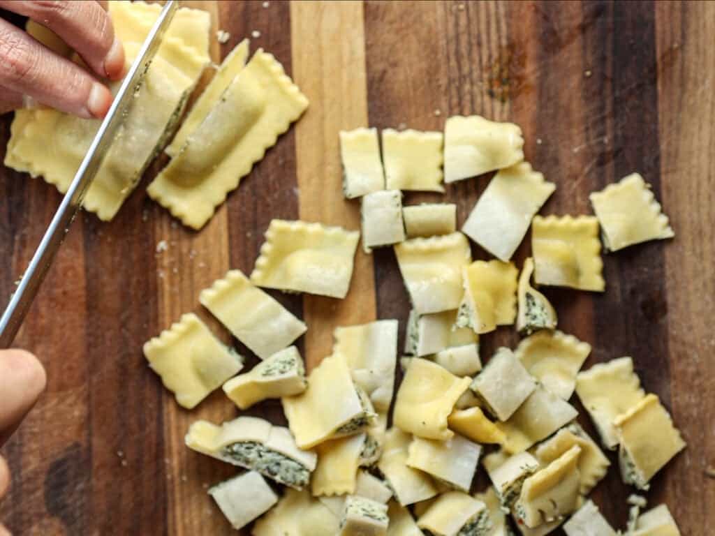 Creamy Cozy Kale White Bean Soup (EASY & Vegan). 14 A hand slices ravioli into smaller pieces on a wooden cutting board, with several chopped pasta pieces scattered around.