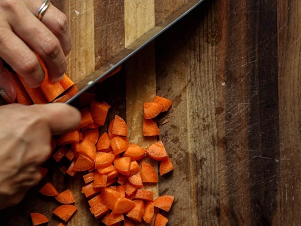 Creamy Cozy Kale White Bean Soup (EASY & Vegan). 6 A close-up of hands slicing fresh carrots on a wooden cutting board with a large knife. The chopped carrot pieces are scattered on the board.
