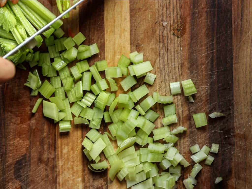 Creamy Cozy Kale White Bean Soup (EASY & Vegan). 7 Chopped celery pieces scattered on a wooden cutting board, with a person’s hand holding a knife, continuing to slice celery stalks.