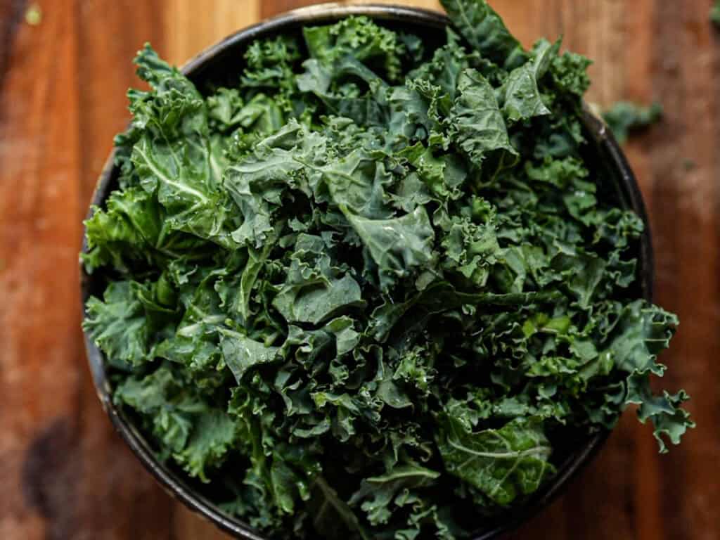 Creamy Cozy Kale White Bean Soup (EASY & Vegan). 4 A bowl filled with fresh, curly green kale leaves sits on a wooden surface.