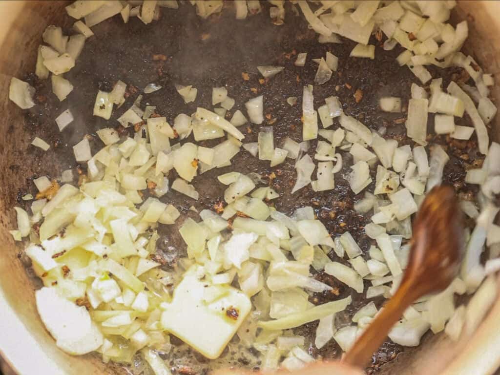 Creamy Cozy Kale White Bean Soup (EASY & Vegan). 16 Chopped onions being sautéed in a pot, turning translucent and lightly browned, with a wooden spoon stirring them.