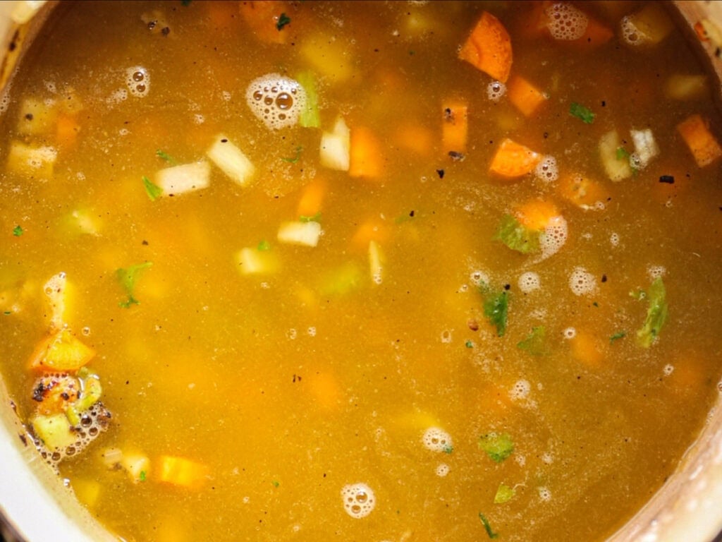 Creamy Cozy Kale White Bean Soup (EASY & Vegan). 20 A close-up of vegetable soup simmering, showing chunks of carrots, celery, and onions in a golden broth with small bubbles and specks of herbs floating on the surface.