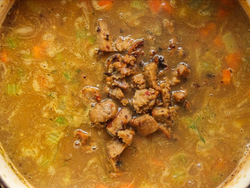 Creamy Cozy Kale White Bean Soup (EASY & Vegan). 21 A close-up of a hearty soup with chopped vegetables and pieces of cooked vegan sausage in a thick, brownish broth. The meat is piled in the center, surrounded by visible celery and carrots.