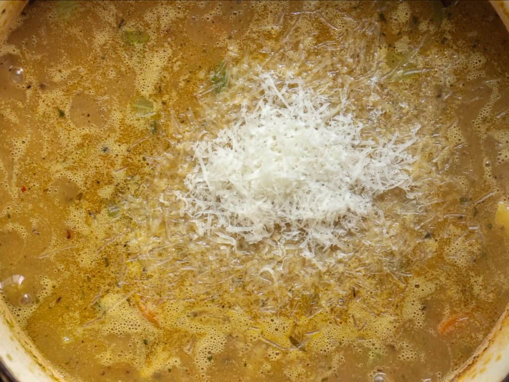 Creamy Cozy Kale White Bean Soup (EASY & Vegan). 11 A close-up of a pot of soup or stew with a heap of grated vegan parmesan melting in the center, surrounded by a creamy, speckled broth and visible vegetable pieces.