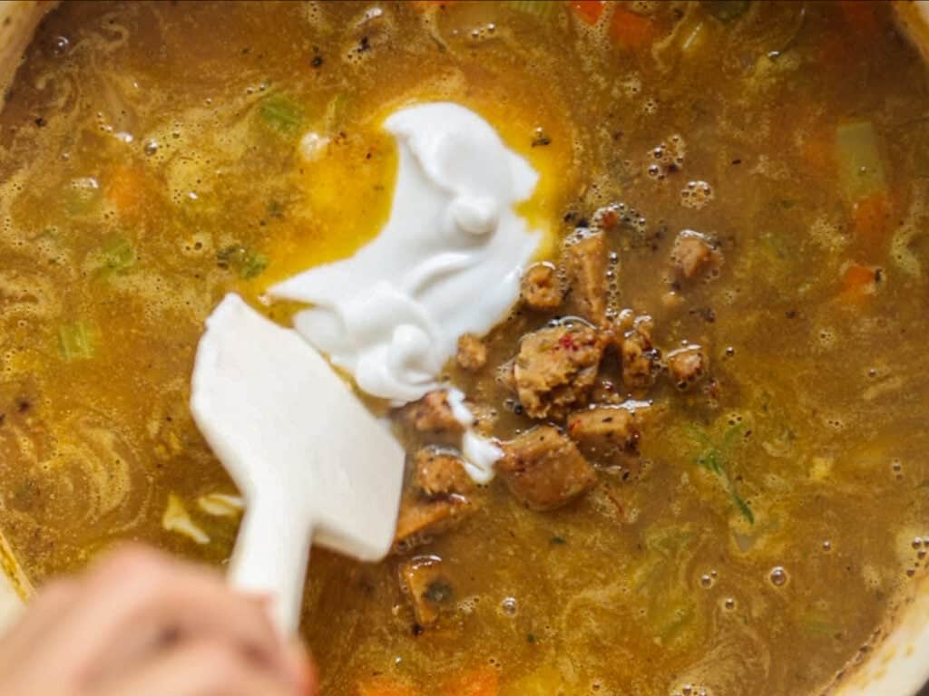 Creamy Cozy Kale White Bean Soup (EASY & Vegan). 23 A close-up of a hand using a white spatula to stir creamy vegan yogurt and chunks of vegan sausage into a pot of brown soup with visible herbs and vegetables.