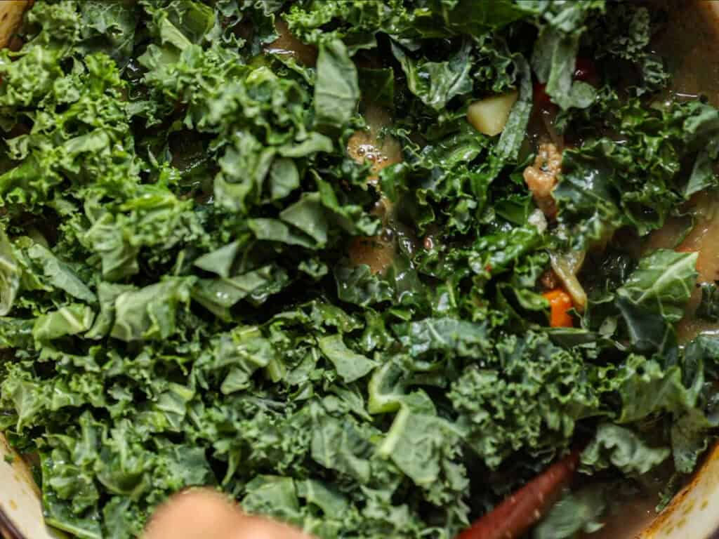Creamy Cozy Kale White Bean Soup (EASY & Vegan). 24 Chopped fresh kale being cooked in a pot with a mixture of vegetables and broth visible underneath.