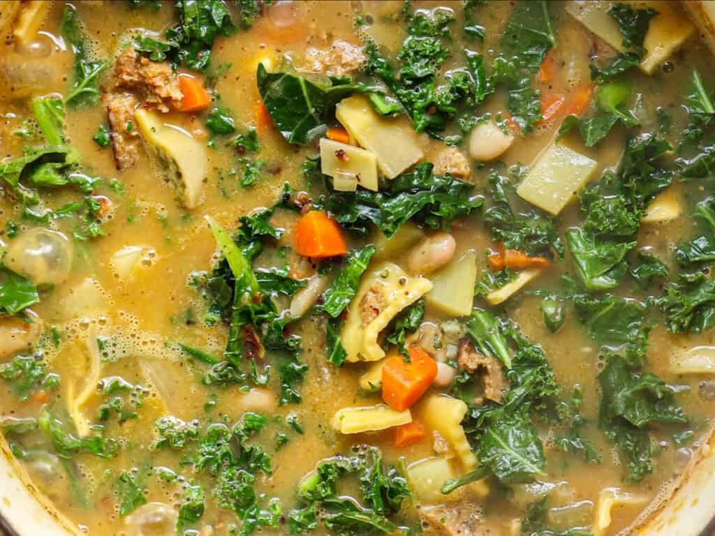 Creamy Cozy Kale White Bean Soup (EASY & Vegan). 2 A close-up of a hearty vegetable soup with kale, carrots, white beans, and chunks of potato in a flavorful broth. The ingredients are vibrant and colorful, giving the soup a fresh, wholesome appearance.