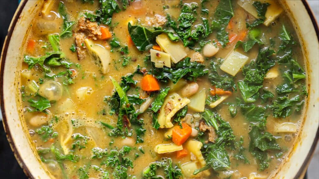 Creamy Cozy Kale White Bean Soup (EASY & Vegan). 25 A close-up of a hearty vegetable kale white bean soup filled with leafy kale, carrots, beans, potatoes, and other vegetables in a light broth, simmering in a pot.