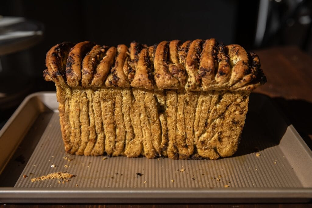 The Korean Vegan: HomeMade 13 A homemade loaf of pull-apart bread with a crispy, golden-brown crust, topped with herbs and spices, sits on a baking tray. The breads layers are visible, creating a textured, accordion-like appearance—a delightful addition to any collection of vegan bread recipes.